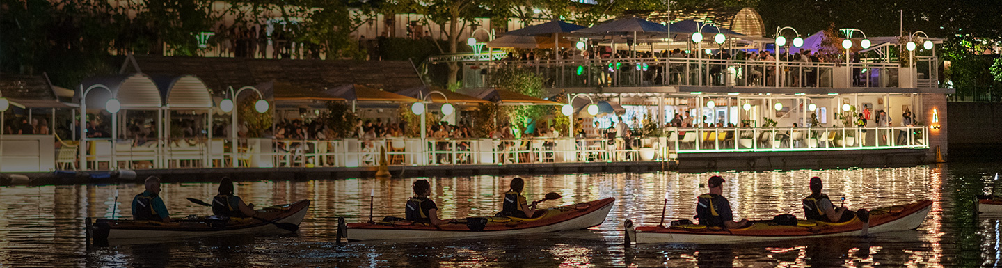 People boating on a river at night with busy restaurant in the background