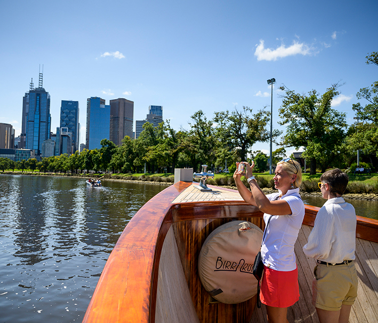 Two middle aged women in a boat on the Yarra River travelling towards Melbourne city, one is taking a photo