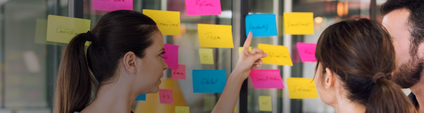 woman looking at a wall covered in square post it notes, discussing plan with male person